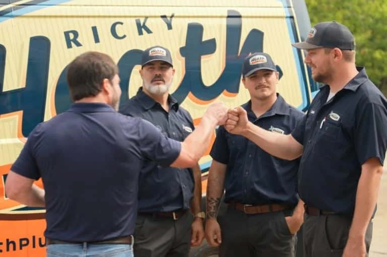 Four Ricky Heath technicians in uniform, two fist-bumping, stand proudly in front of a service van.